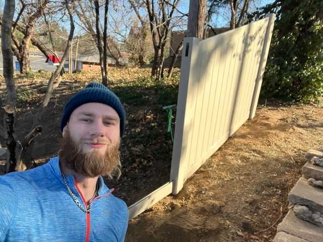 Selfie beside a pale yellow fence panel in a wooded yard, with stone steps and a red hammock in the background