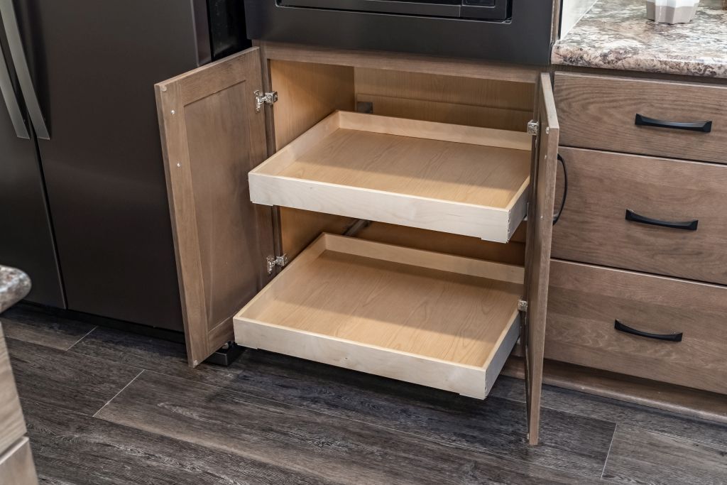 Kitchen cabinet with pull-out shelves, open. Light wood, neutral tones, countertop visible on the right, dark appliances.