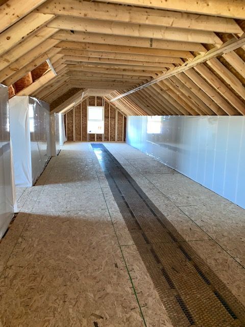Interior of an unfinished attic with wood beams, plywood floor, and windows.