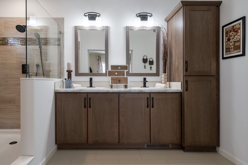 Bathroom with double vanity, mirrors, shower, and tall storage cabinet. Brown cabinets, white countertops, and neutral walls.