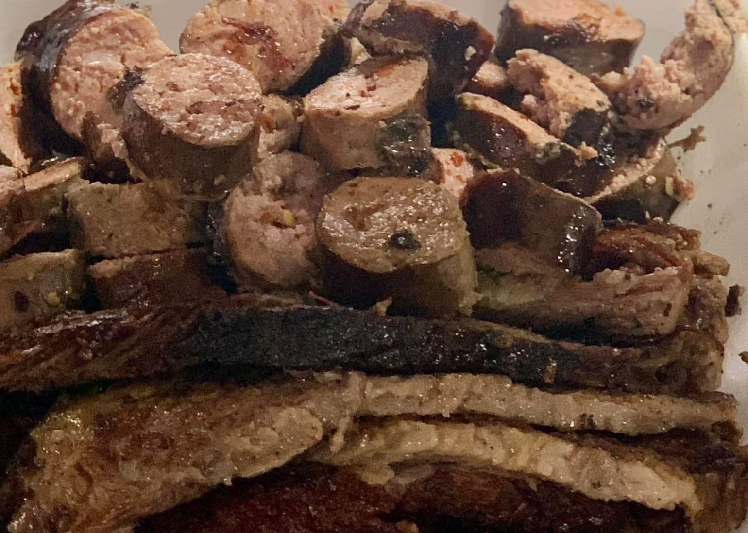 A bowl containing a variety of sliced, cooked barbecue meats, including dark-crusted brisket and rounds of sausage.