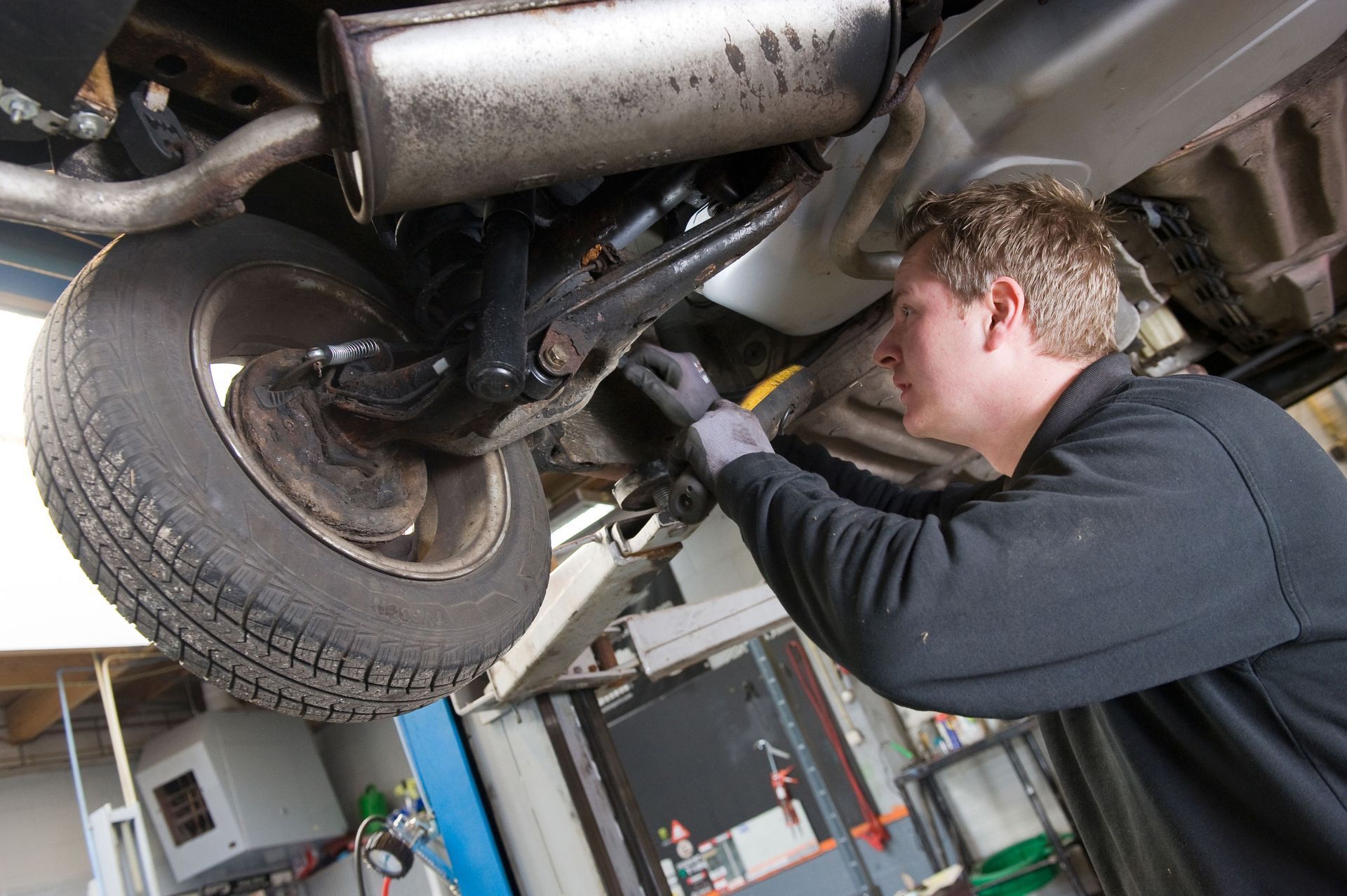 Mechanic working on the underside of a car in a garage, wearing gloves.