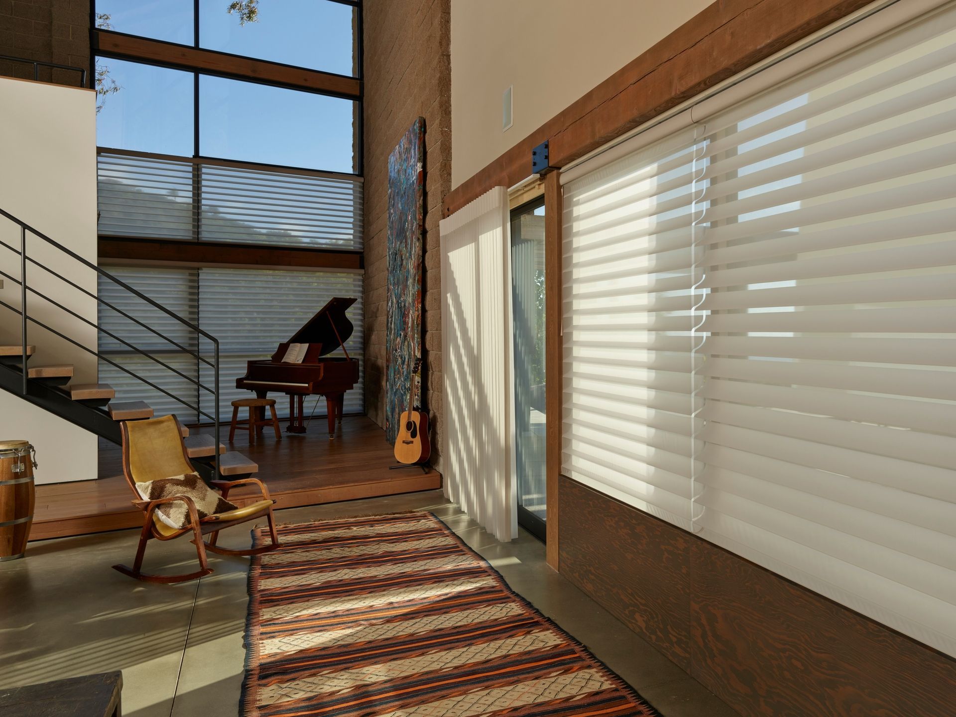Modern living room with staircase, grand piano, rocking chair, and sunlit windows with blinds
