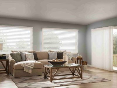 A bright, neutral-toned living room featuring a tan sectional sofa, a wooden coffee table, and large windows with blinds.