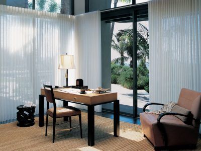 A home office with a modern desk, chair, and armchair, set before floor-to-ceiling white curtains overlooking a patio.