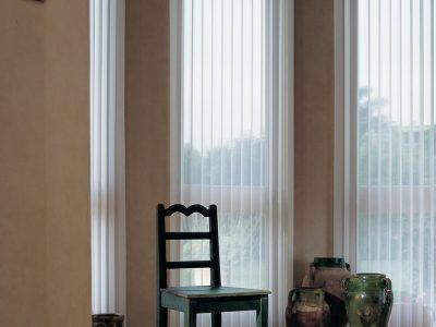 A wooden chair sits in a bright room in front of large windows with sheer white vertical blinds and several ceramic vases.