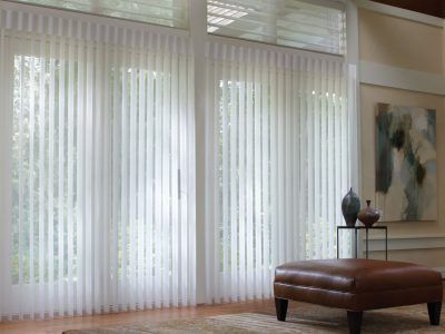 Sheer white vertical blinds cover large windows next to a brown leather ottoman and a framed painting on a beige wall.