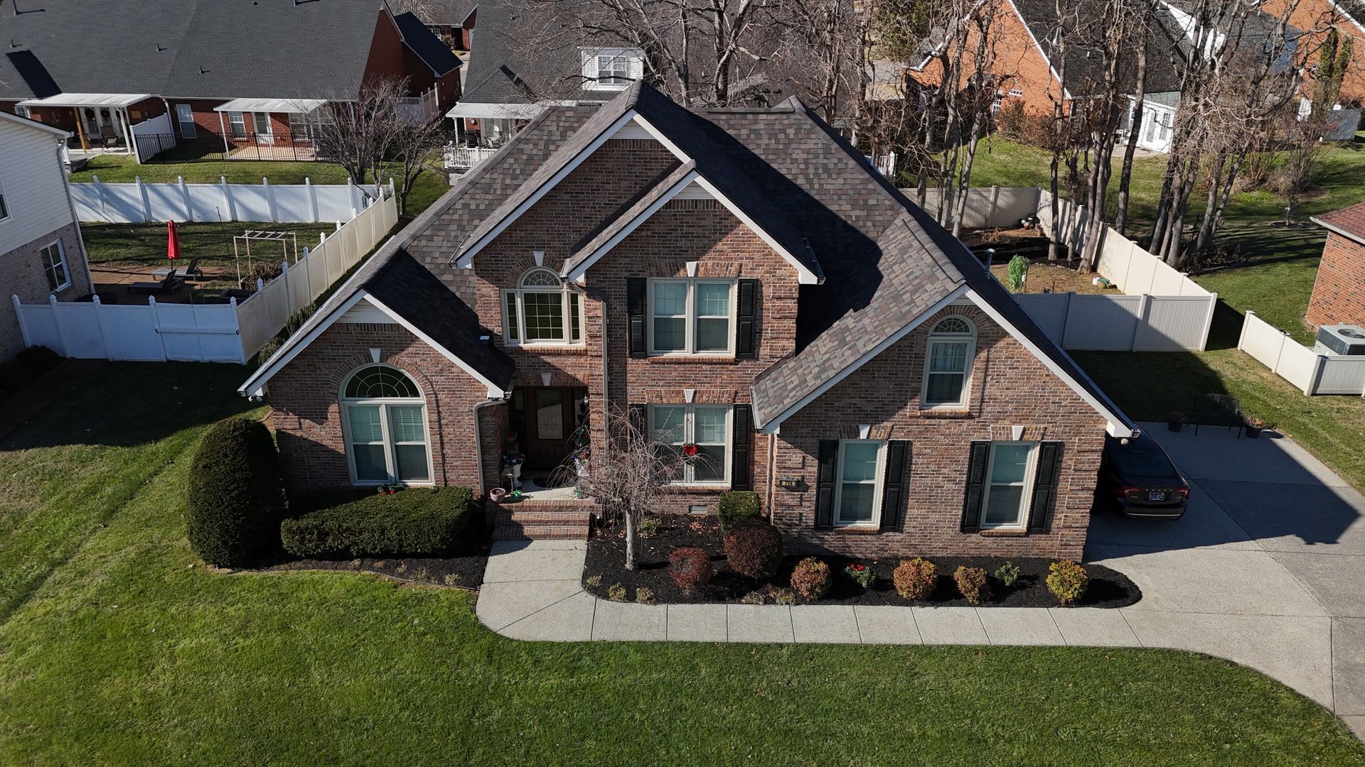 Aerial view of a two-story brick suburban house with a dark shingled roof, front lawn, and concrete driveway.