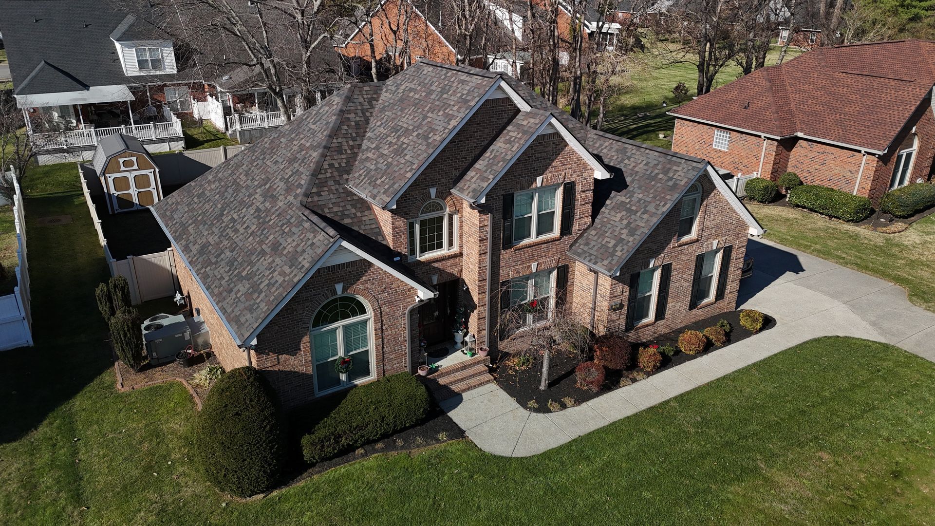 An aerial view of a two-story brick suburban house with a gray shingled roof on a sunny day.