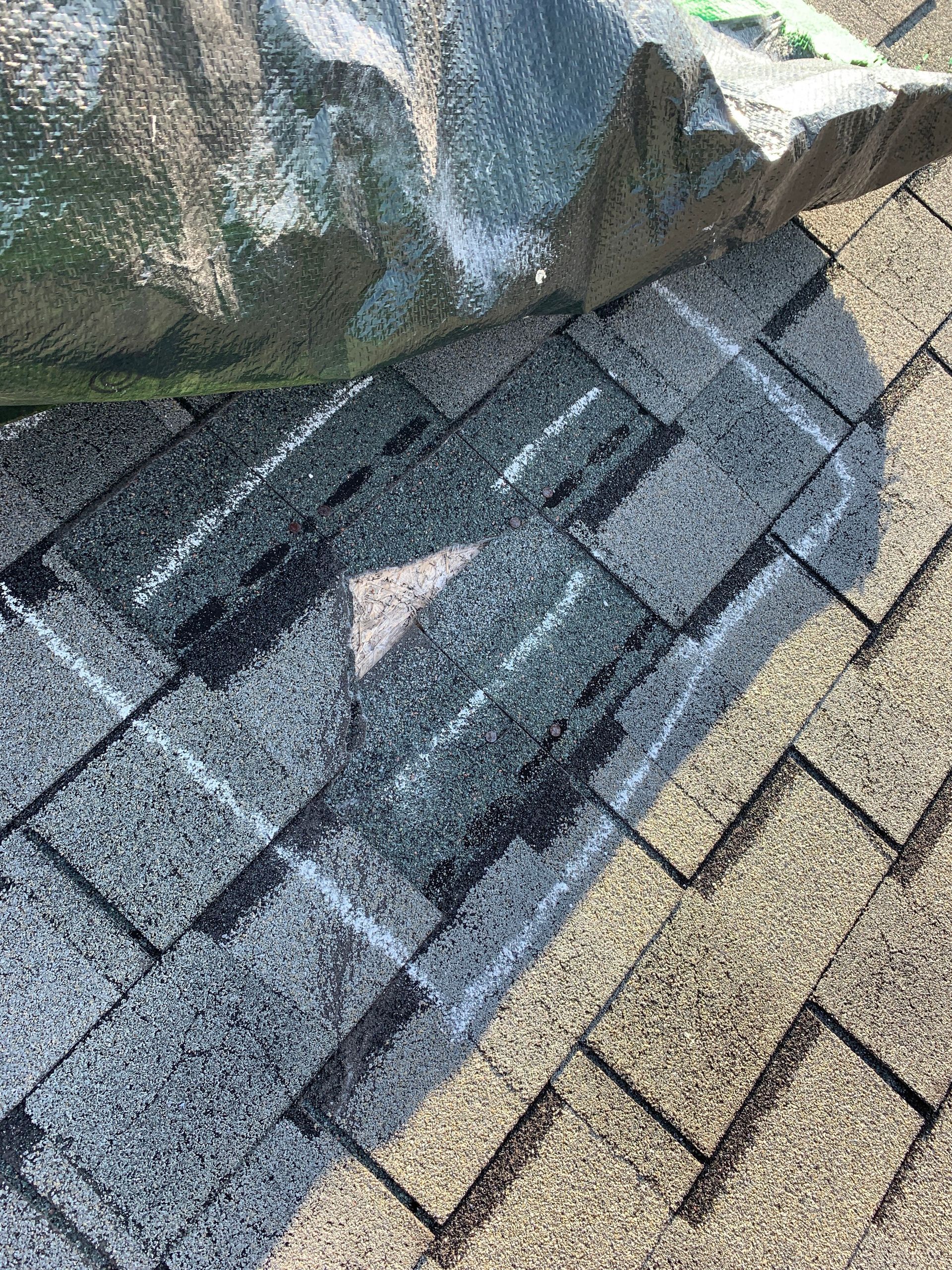 A large piece of rock sits on a residential roof, crushing and damaging several gray asphalt shingles.