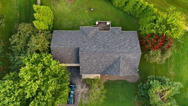 An aerial view of a gray-roofed house surrounded by green grass and trees in a residential setting.