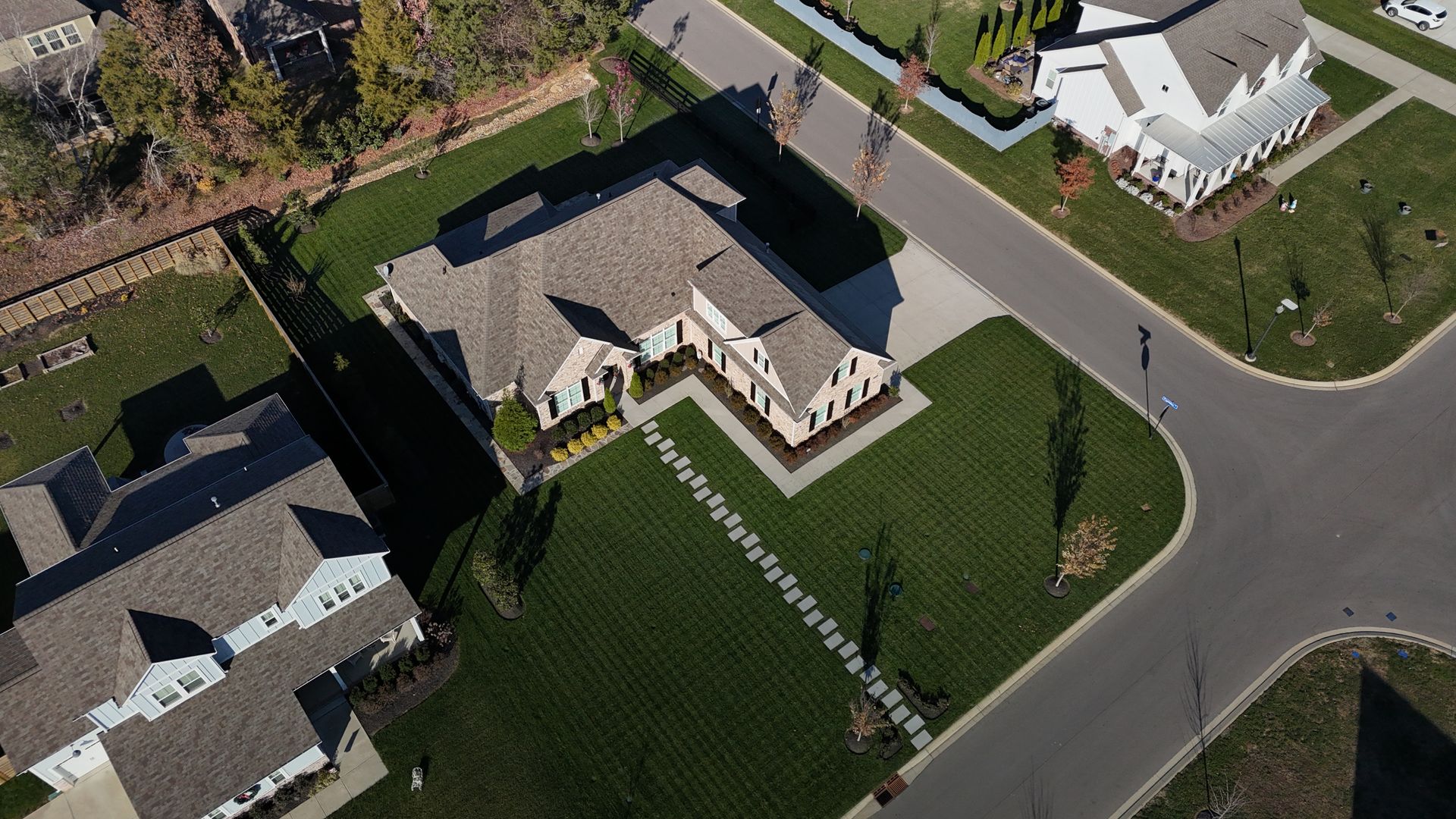 An aerial view shows a suburban neighborhood with houses, green lawns, and a paved street corner on a sunny day.