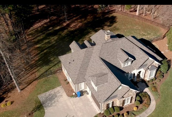 Aerial view of a large residential home with a grey shingle roof, stone siding, a concrete driveway, and a grassy yard.
