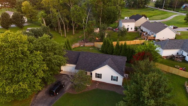 Aerial view of a white single-story suburban house with a dark gray roof, surrounded by trees and neighboring homes.