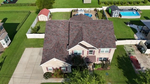Aerial view of a two-story residential house with a dark shingled roof, surrounding lawn, shed, and a backyard pool.