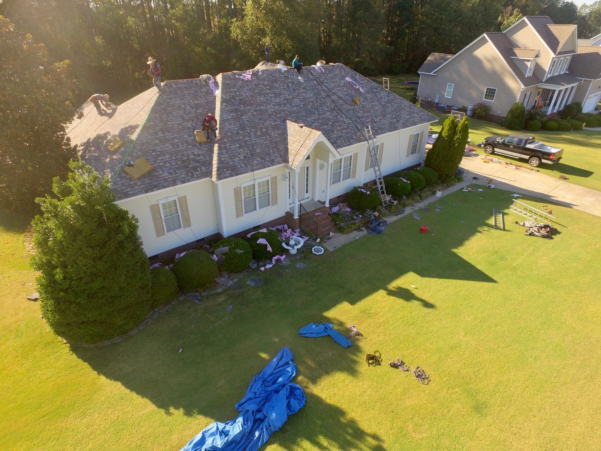 An aerial view of a single-story house undergoing a roof replacement with workers on the shingles and debris below.