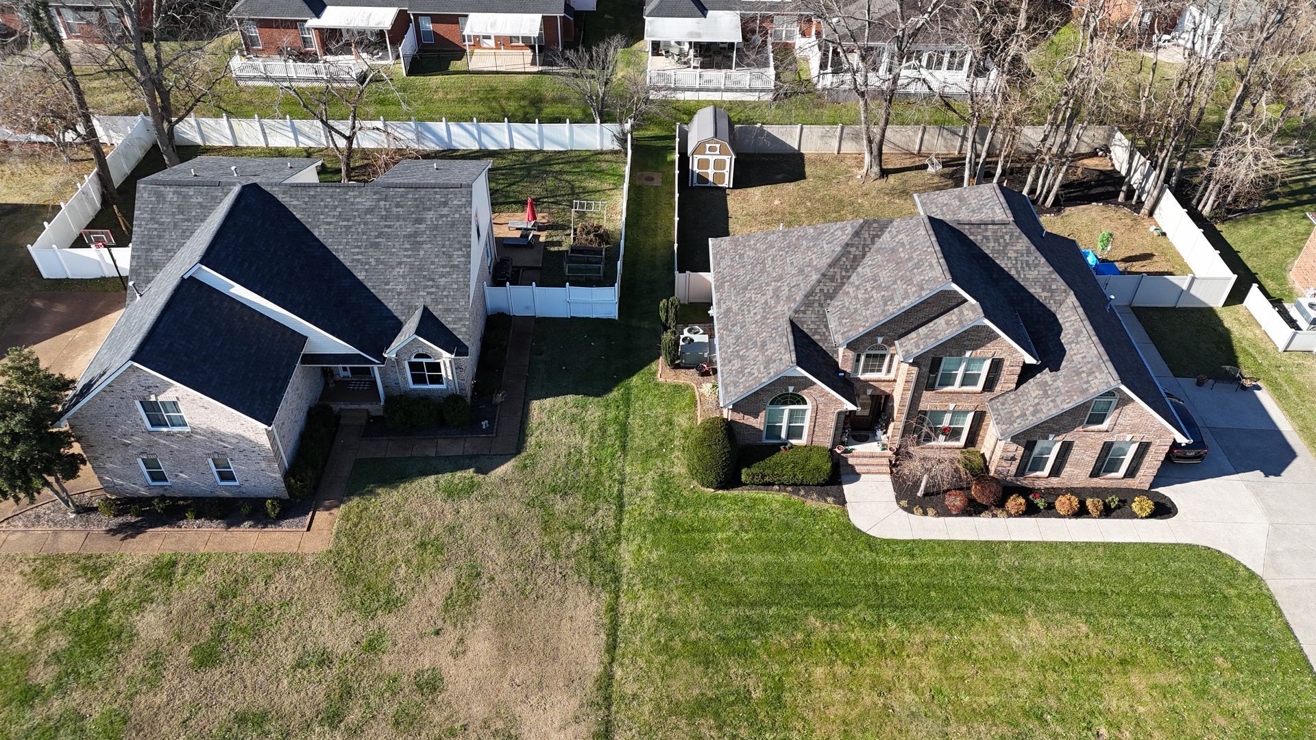 An aerial view of two adjacent brick houses with shingled roofs, separated by a white fence and a narrow patch of grass.
