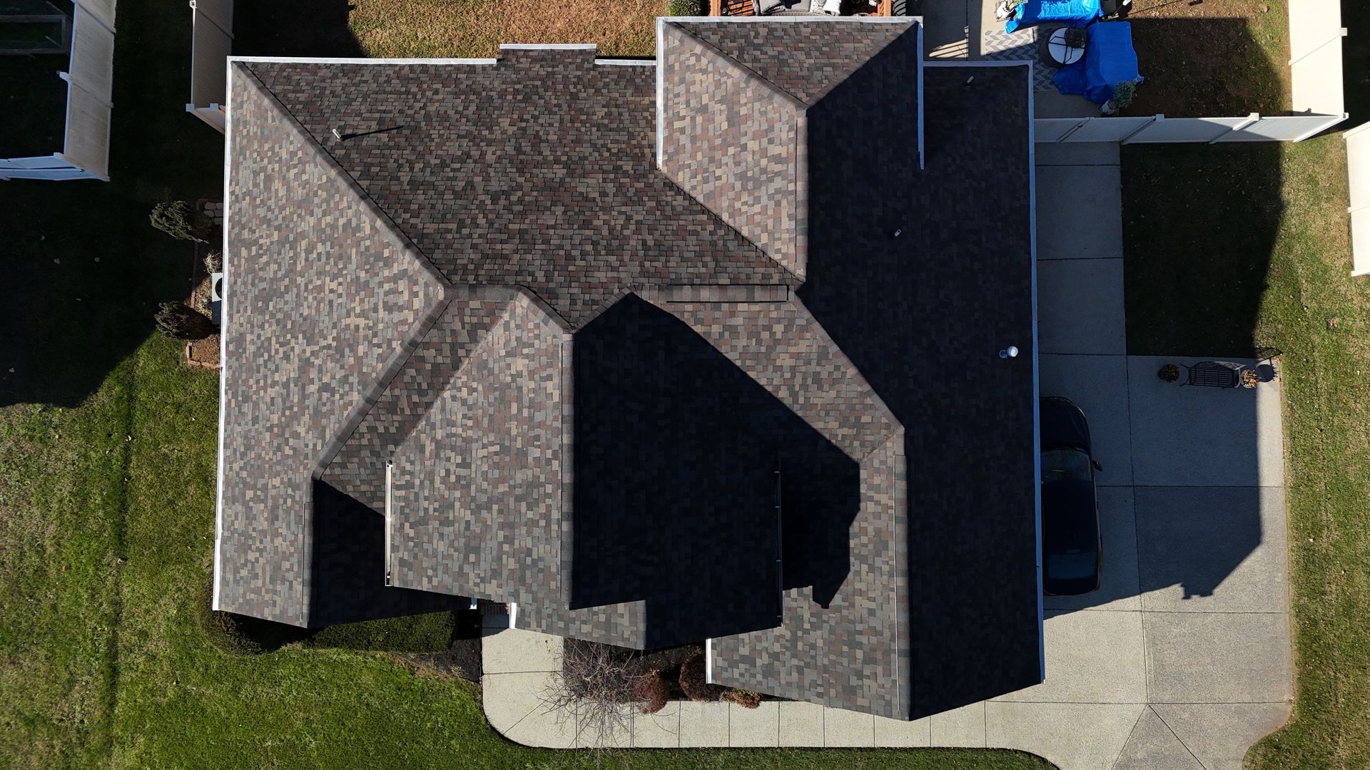 A top-down aerial view of a dark shingled residential roof, showing multiple gables and a driveway beside a grassy lawn.