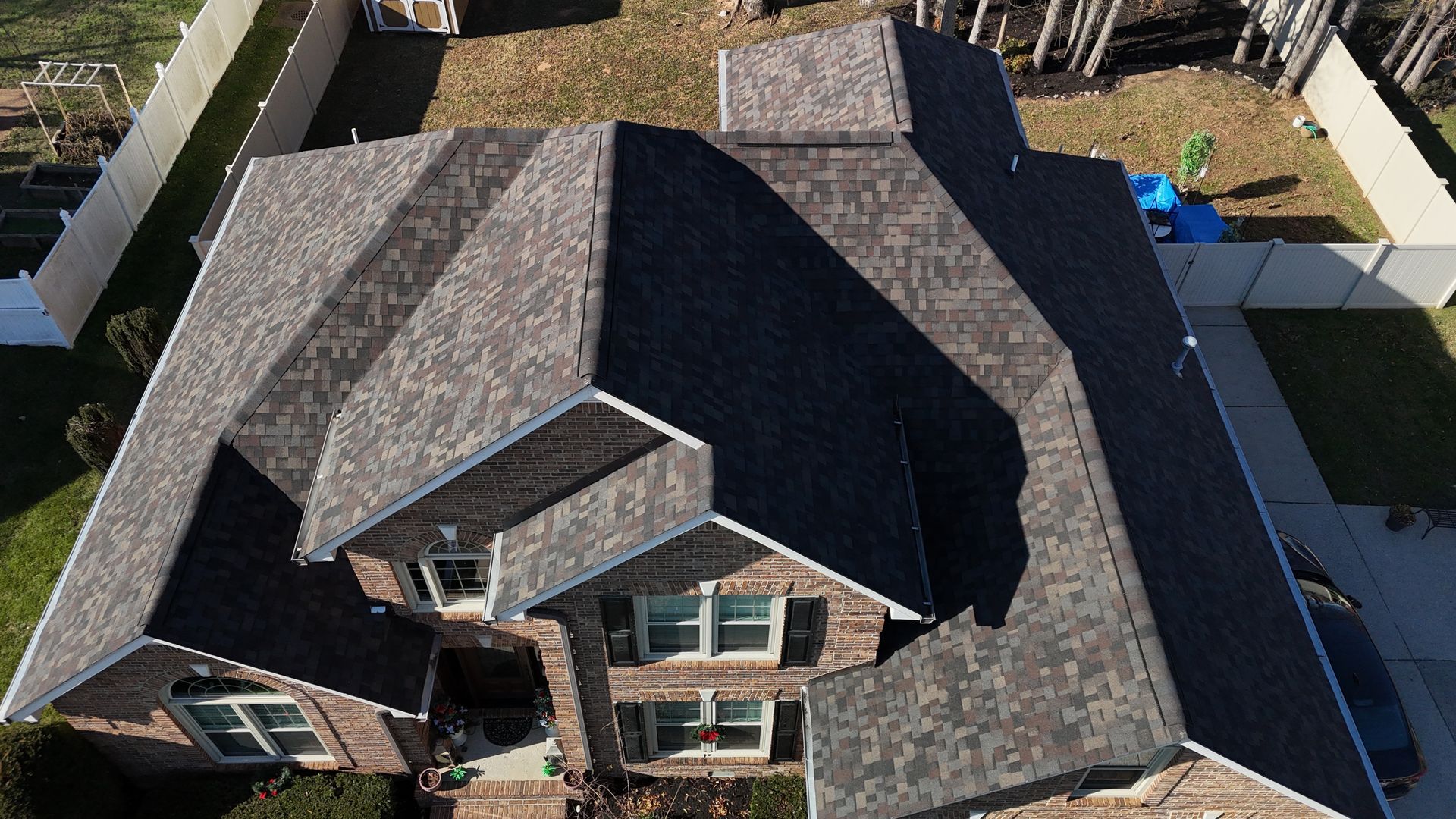 Aerial view of a residential house with a dark shingled roof, brown brick exterior, and a fenced backyard.