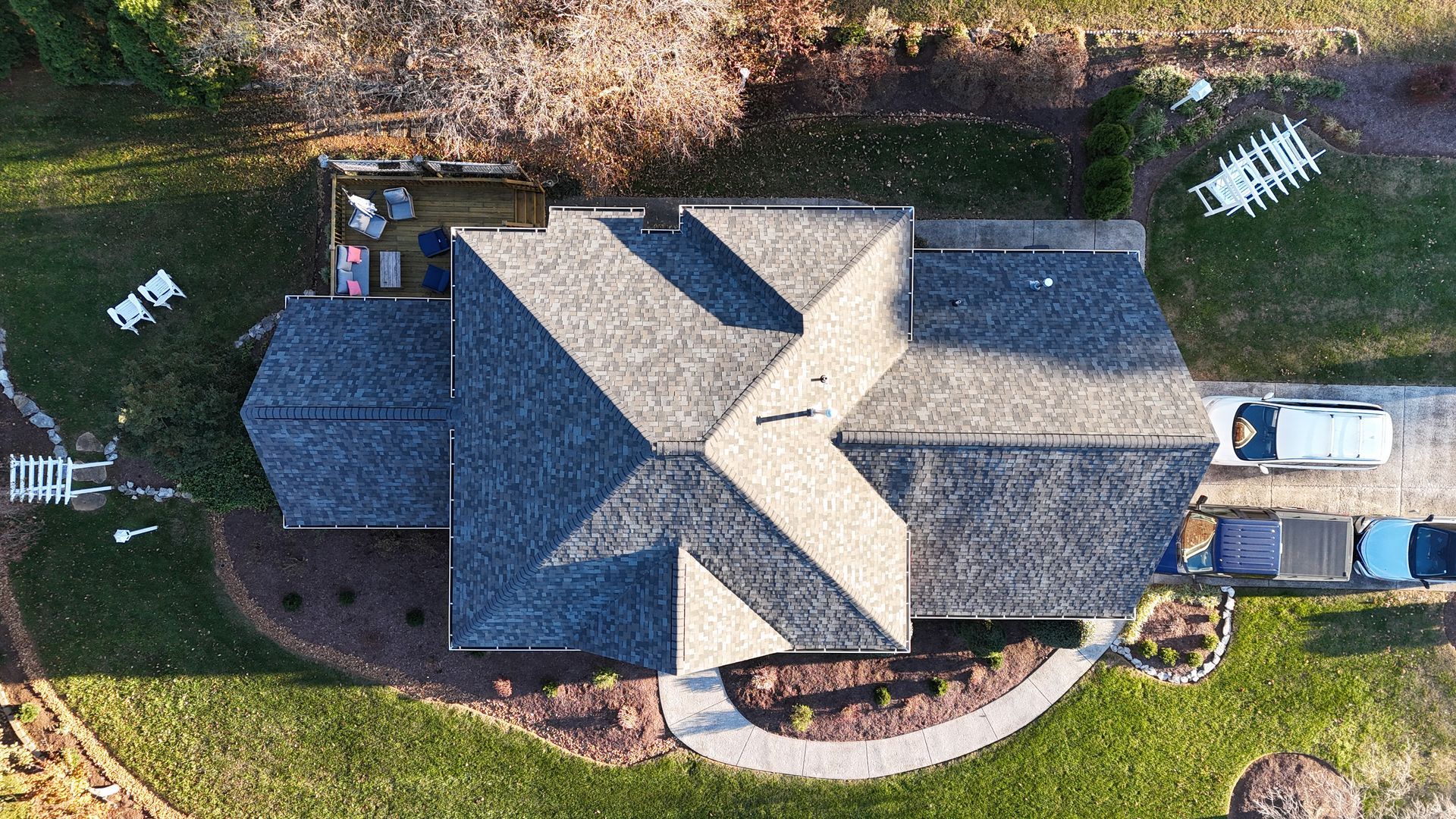 An aerial, top-down view of a house with a gray shingled roof, surrounded by a lawn, garden beds, and a driveway.