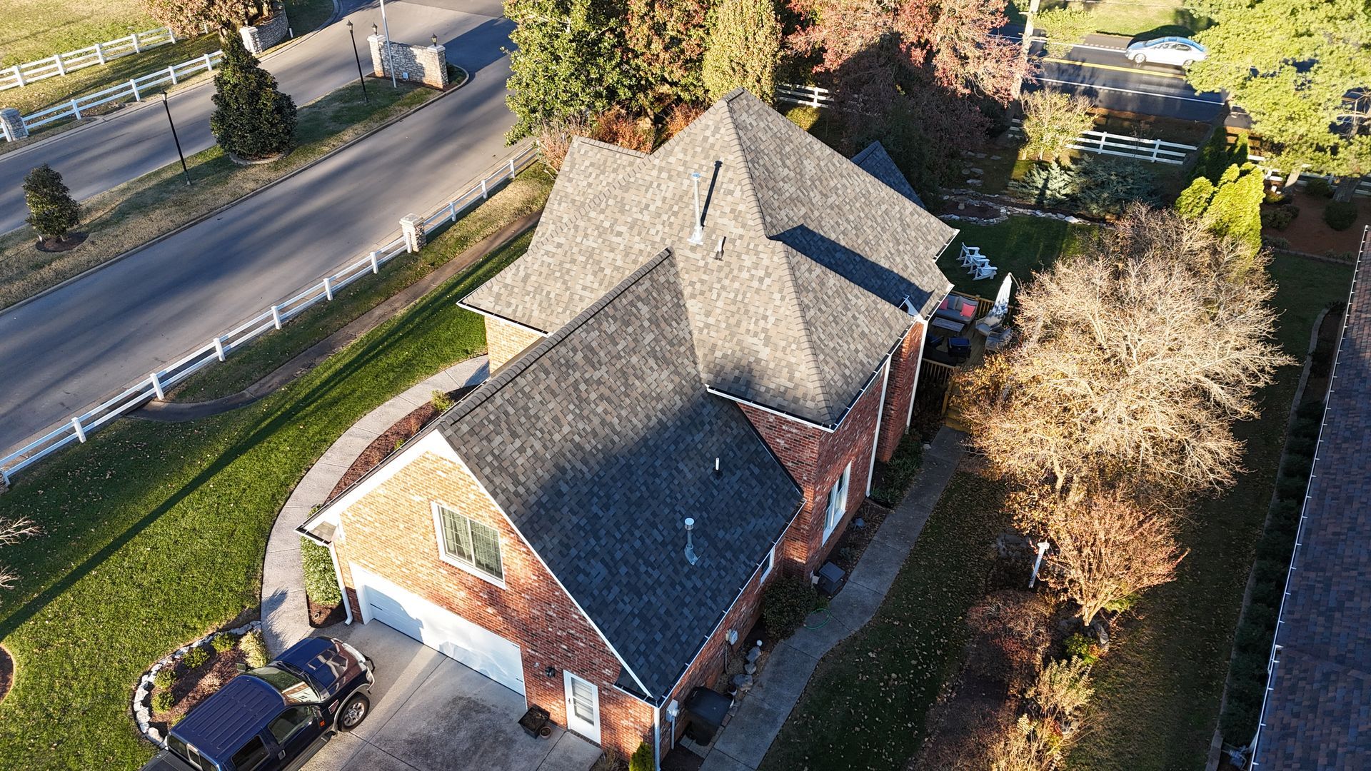 An aerial view of a multi-story brick house with a gray shingled roof, a driveway with a car, and surrounding trees.