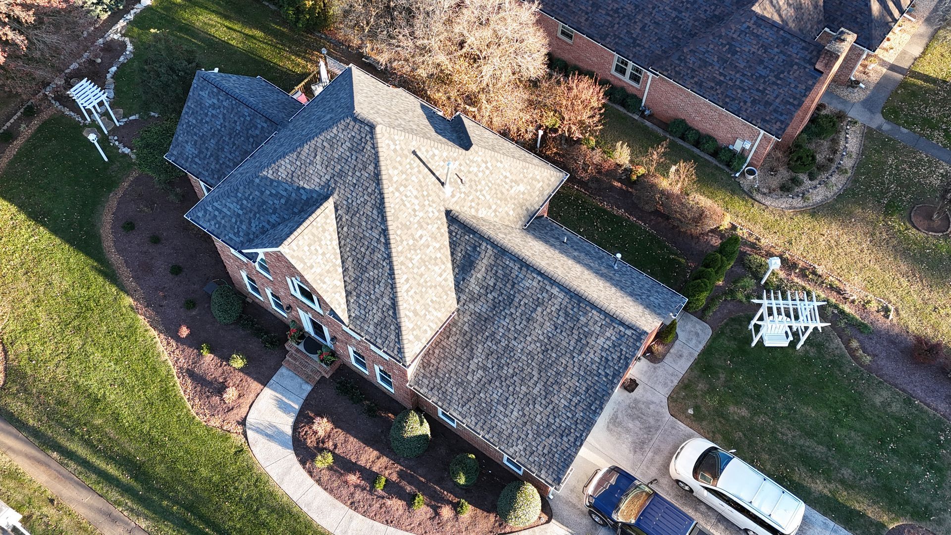 Aerial view of a residential house with a dark shingled roof, surrounding lawn, concrete walkway, and a white vehicle.