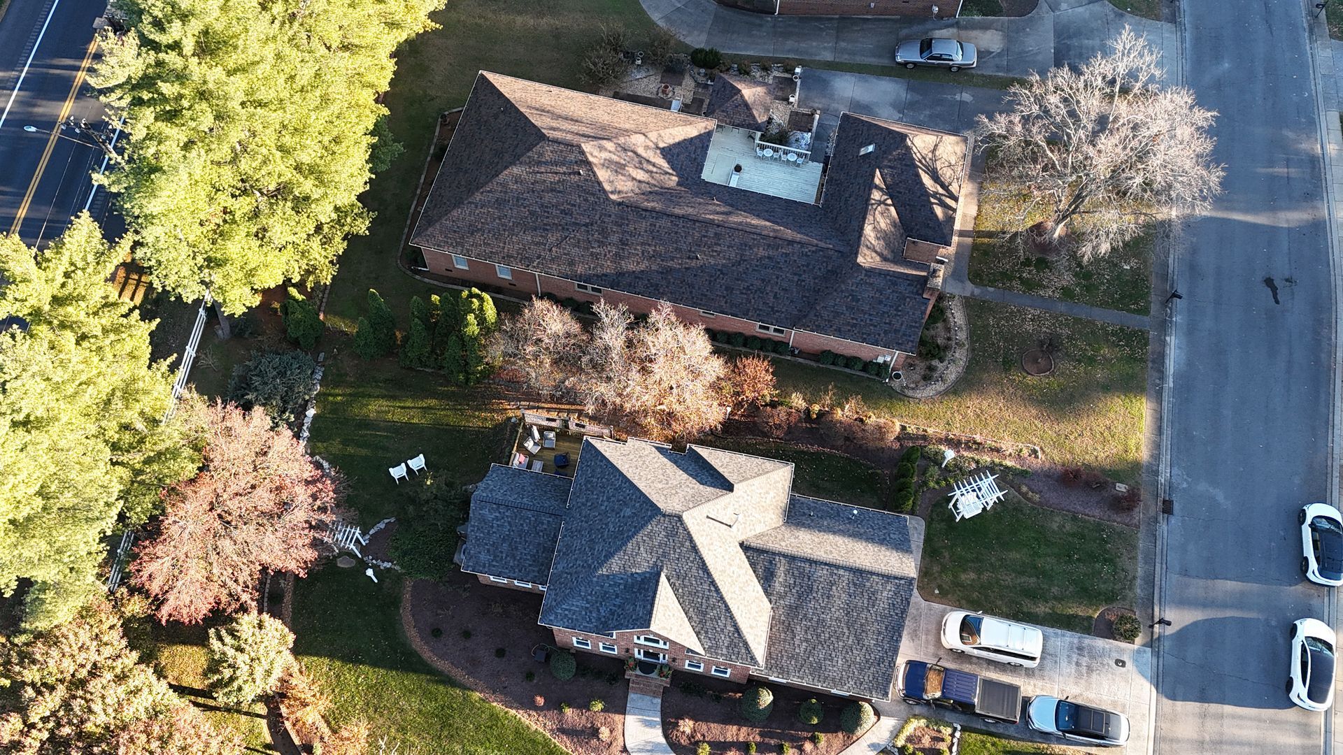 An aerial view of two neighboring residential houses with dark shingled roofs, surrounded by trees and a paved street.