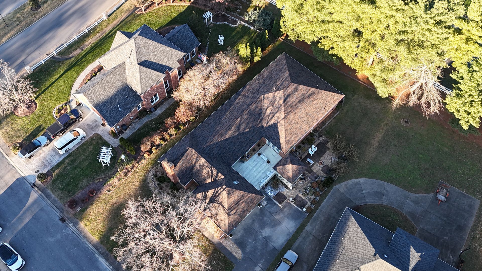 An aerial view of three residential homes with gray shingled roofs, surrounding driveways, and scattered trees.