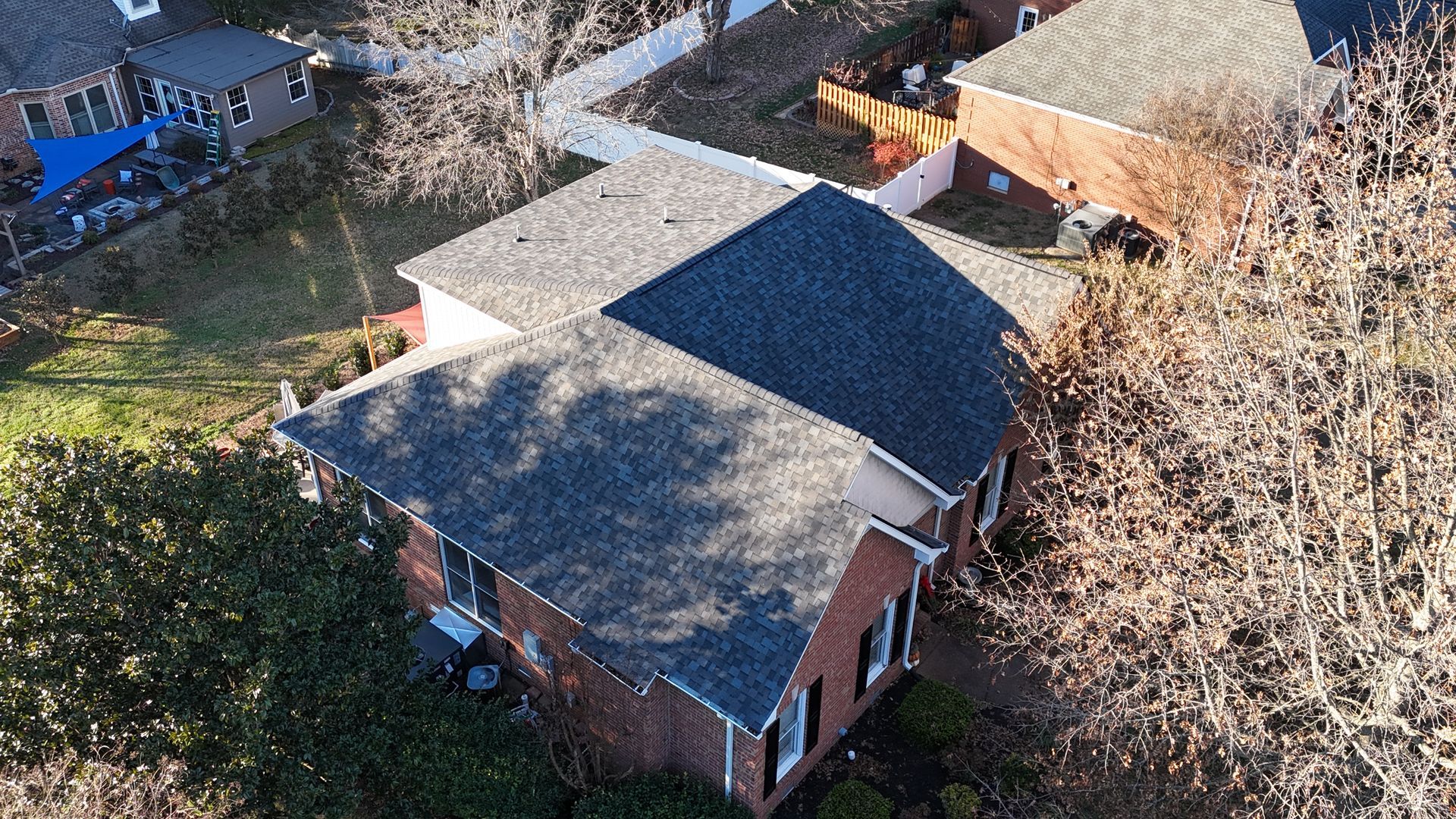An aerial view of a single-story brick residential house with a shingled roof, surrounded by trees and a paved driveway.