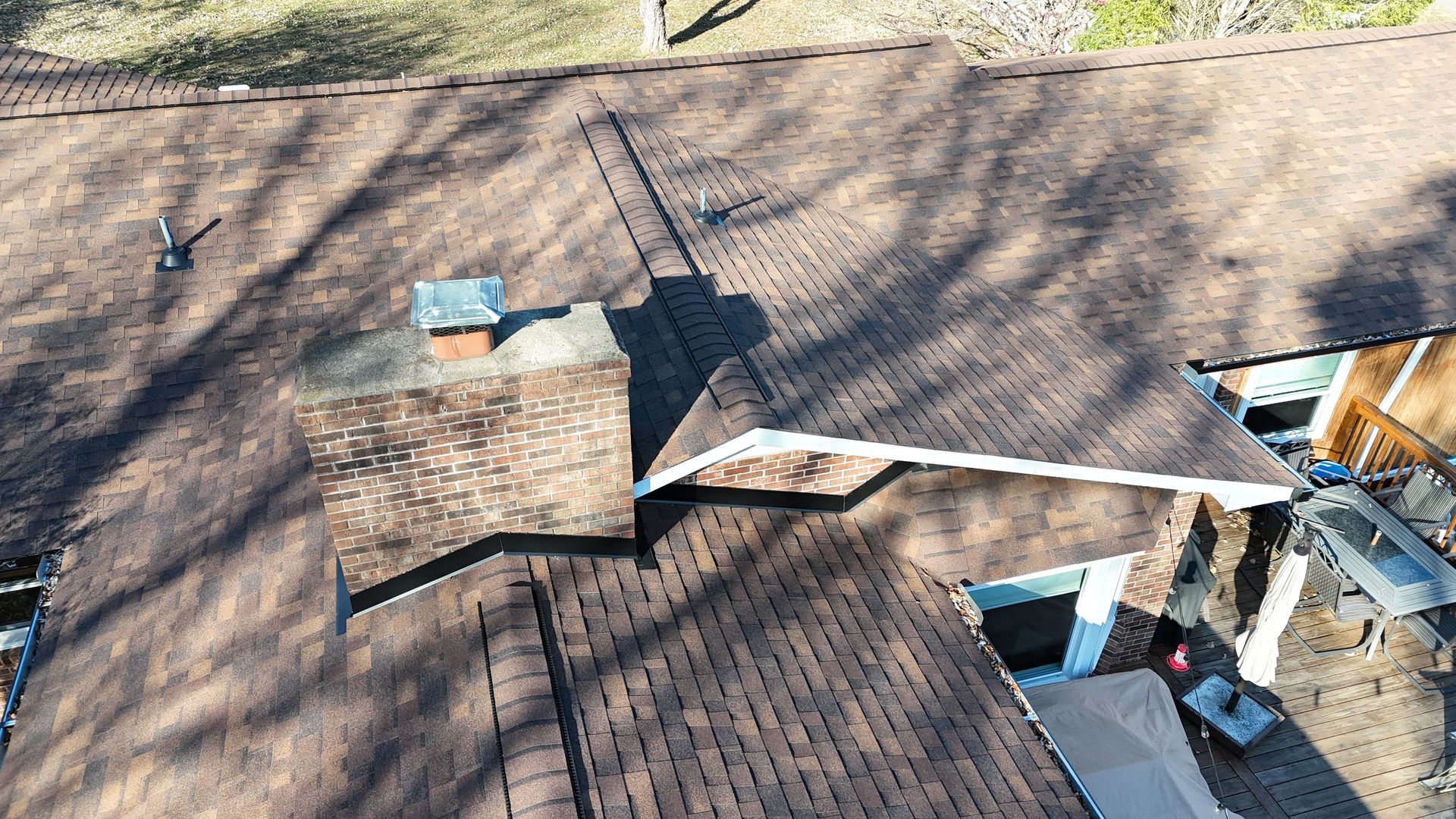 An aerial view of a dark-shingled residential roof with a brick chimney and a covered patio area below.