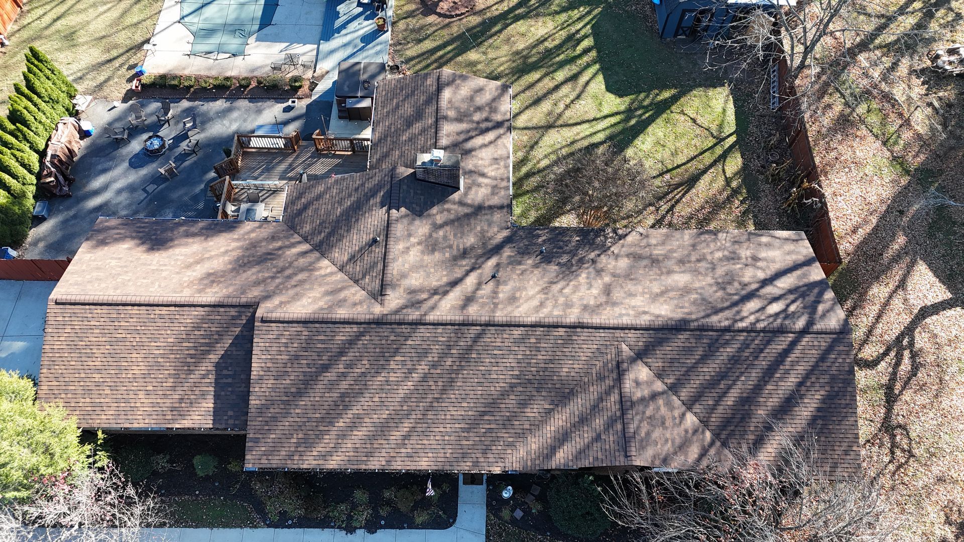 An aerial, top-down view of a brown, shingled residential roof with a chimney, surrounded by a lawn and patio area.