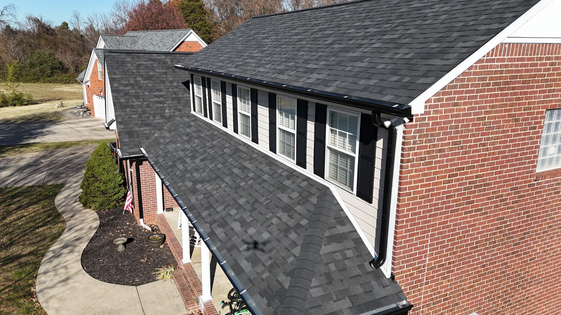 An aerial view of a brick house featuring a dark shingled roof with a dormer and black shutters.
