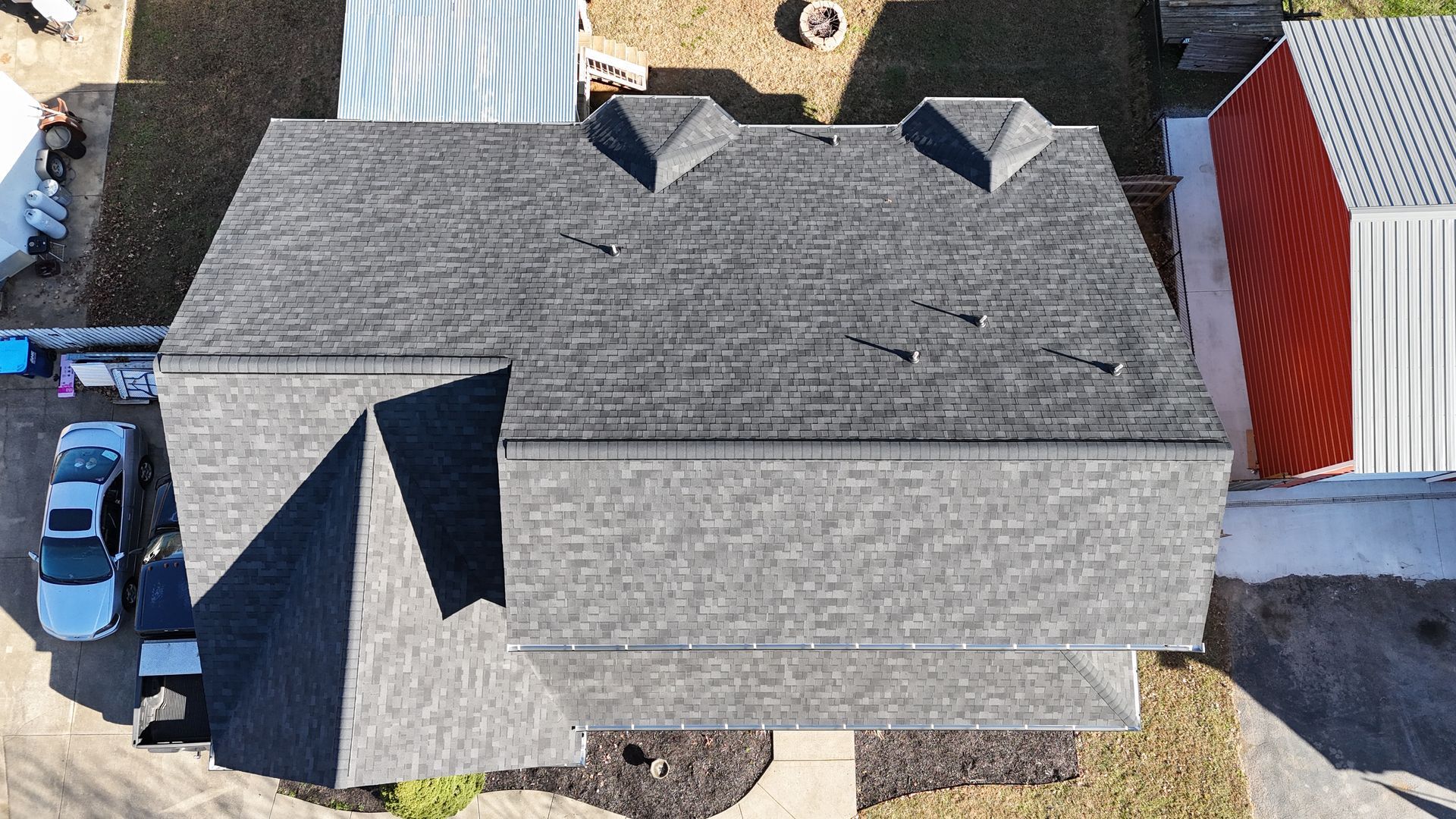 An aerial view of a gray shingled roof with two small skylights, featuring a multi-level design and a partial overhang.