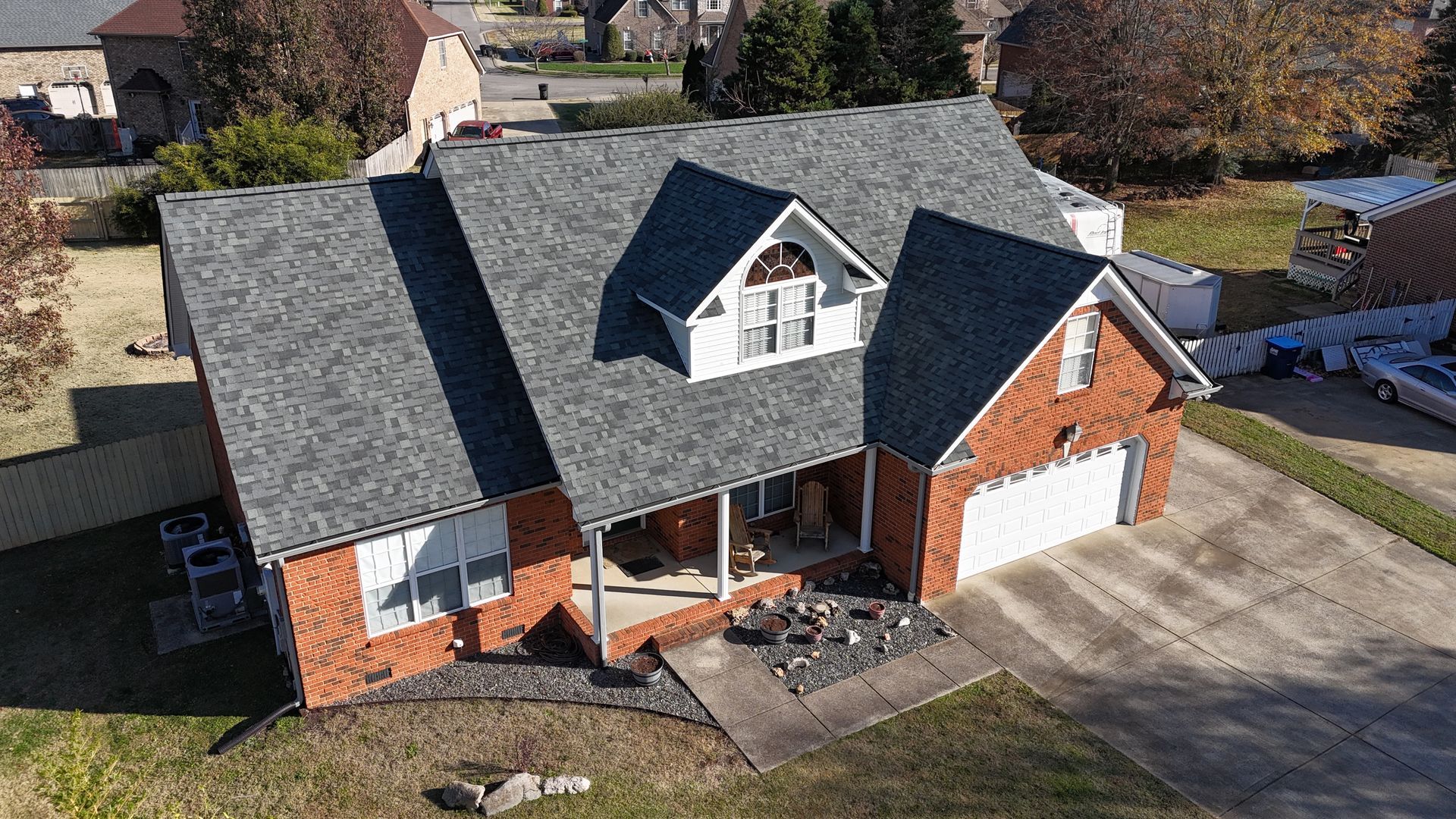 Aerial view of a red brick house with a dark shingled roof, a front porch, and a two-car garage on a sunny day.