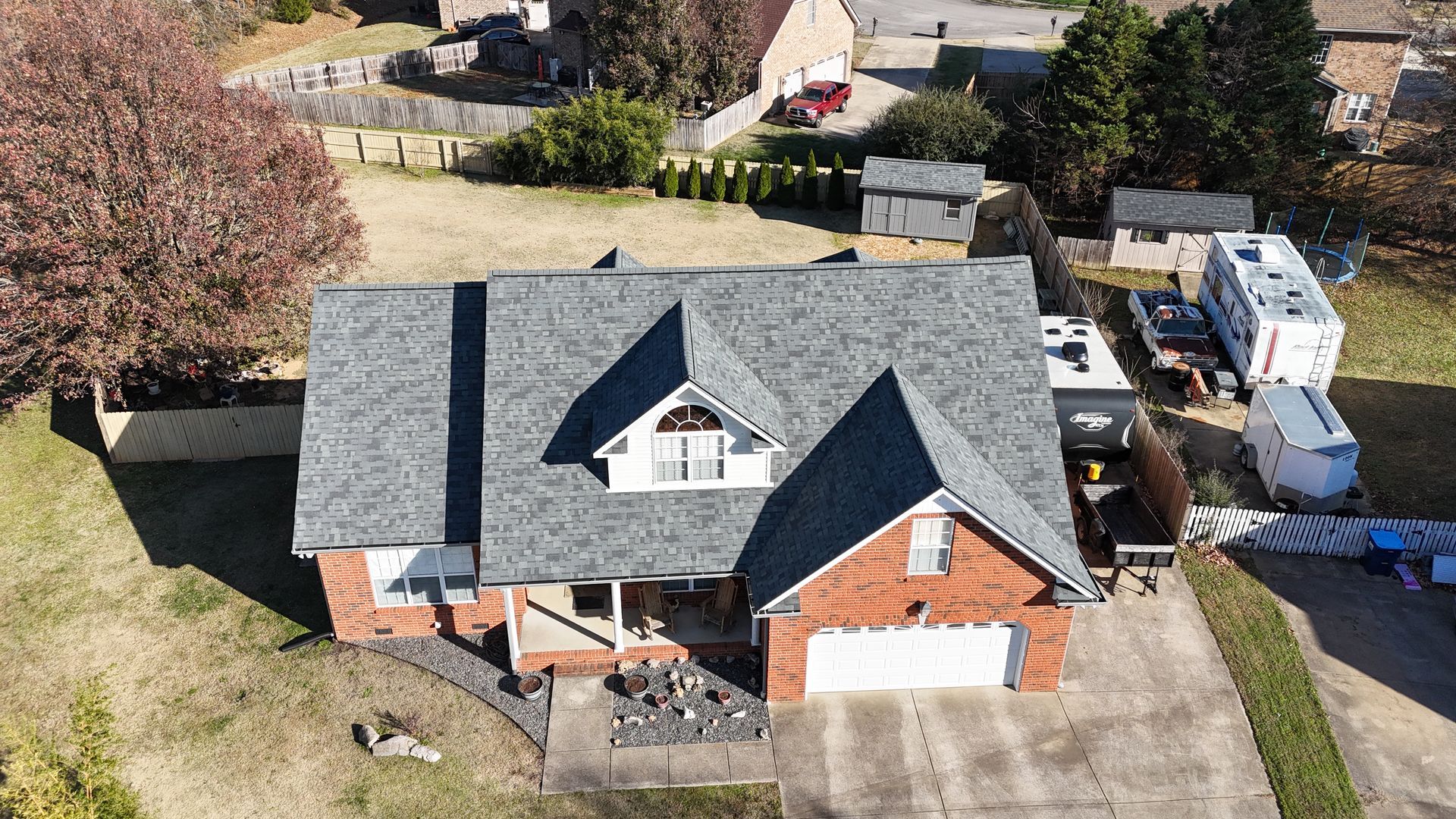 An aerial view of a single-story brick house with a gray shingled roof, a front porch, and an attached two-car garage.