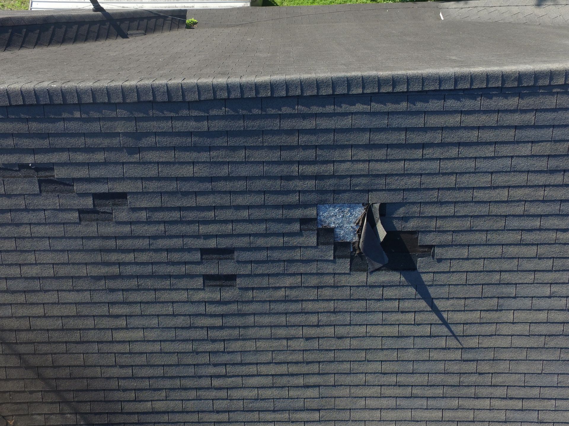 A close-up of a dark grey shingled roof showing localized damage with torn and missing shingles.