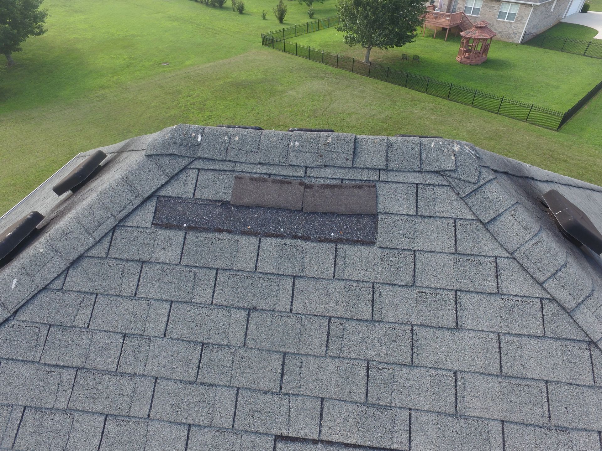A high-angle view of a gray asphalt shingle roof featuring missing shingles and exposed underlayment near the ridge.