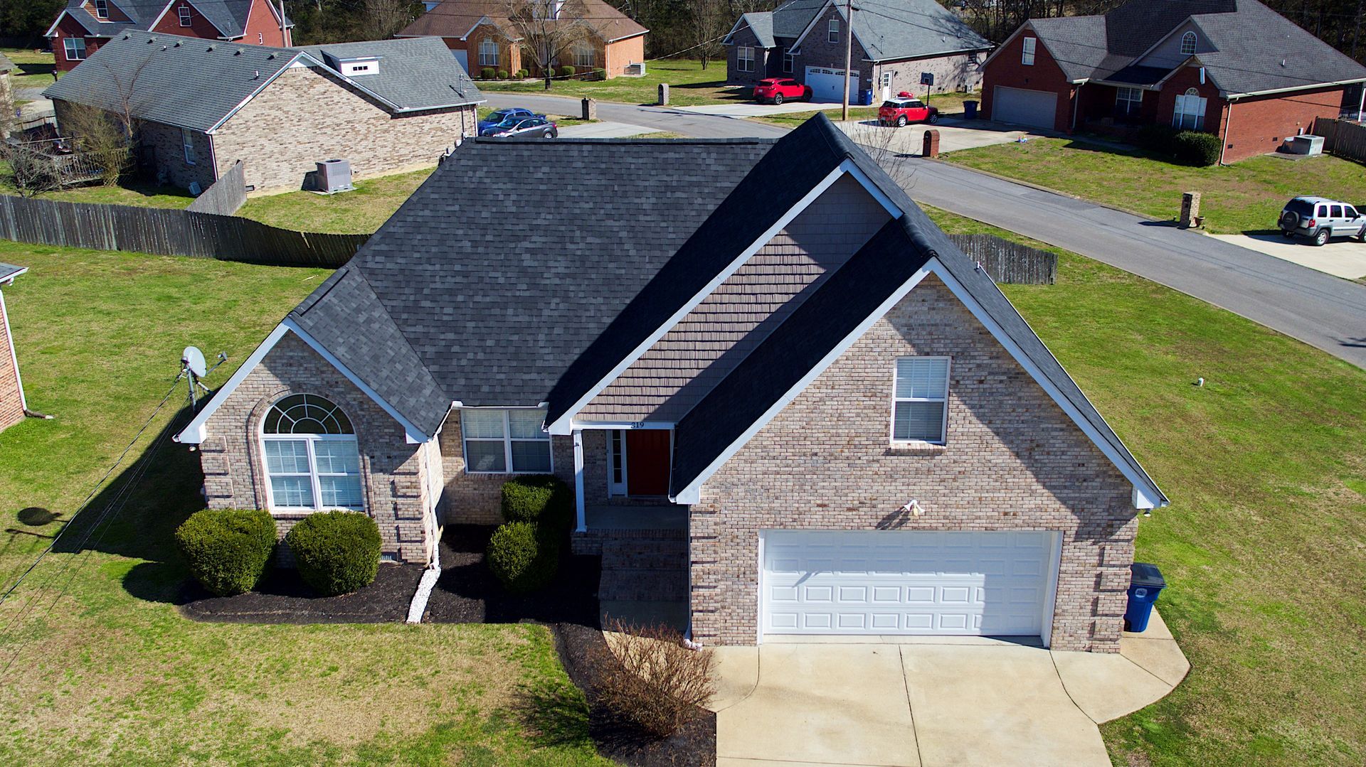 A drone view of a brick house with a dark shingled roof, a front-facing garage, and a lawn in a residential neighborhood.