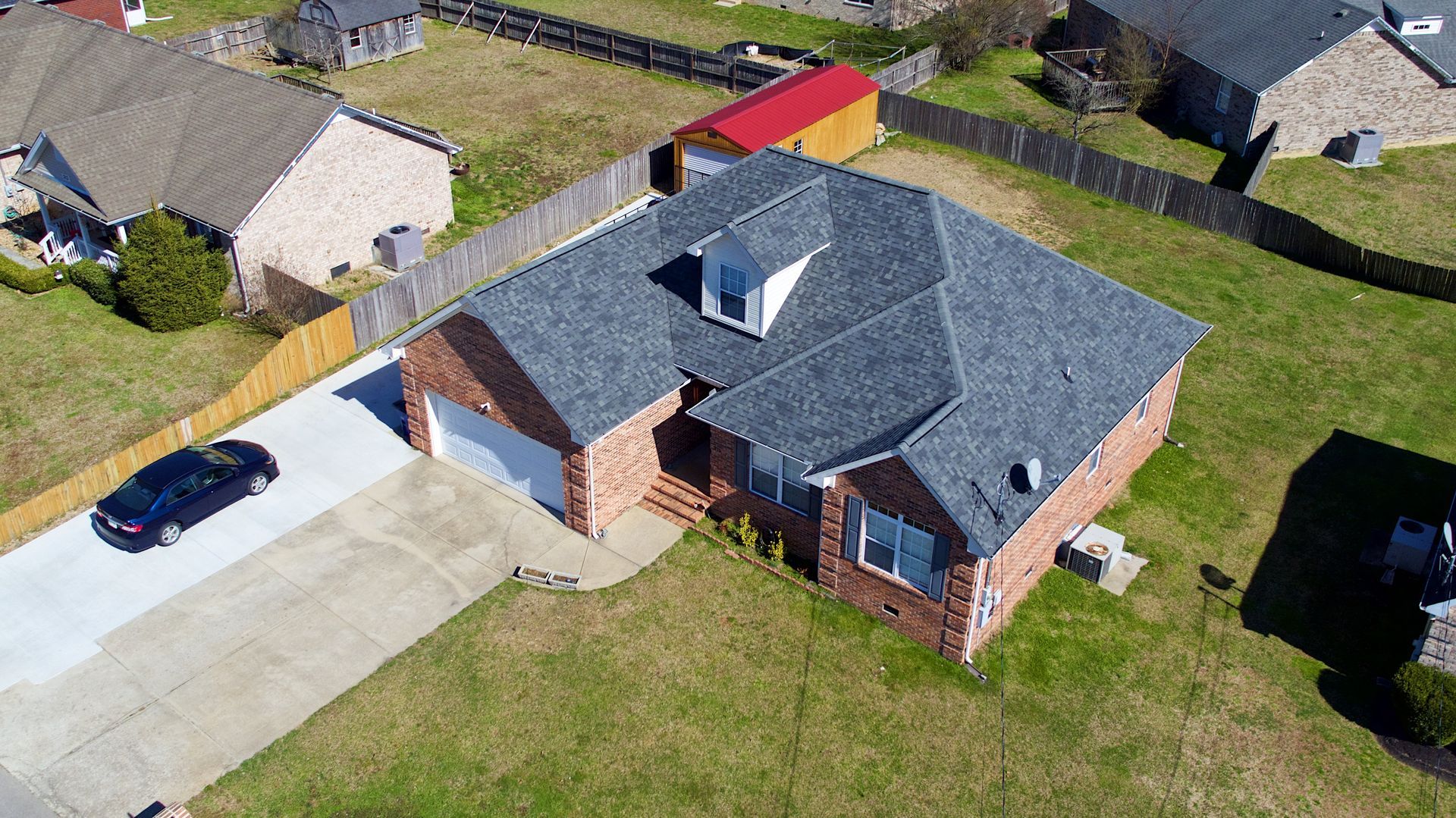 An aerial view of a brick house with a gray roof and a dark sedan parked on the concrete driveway.