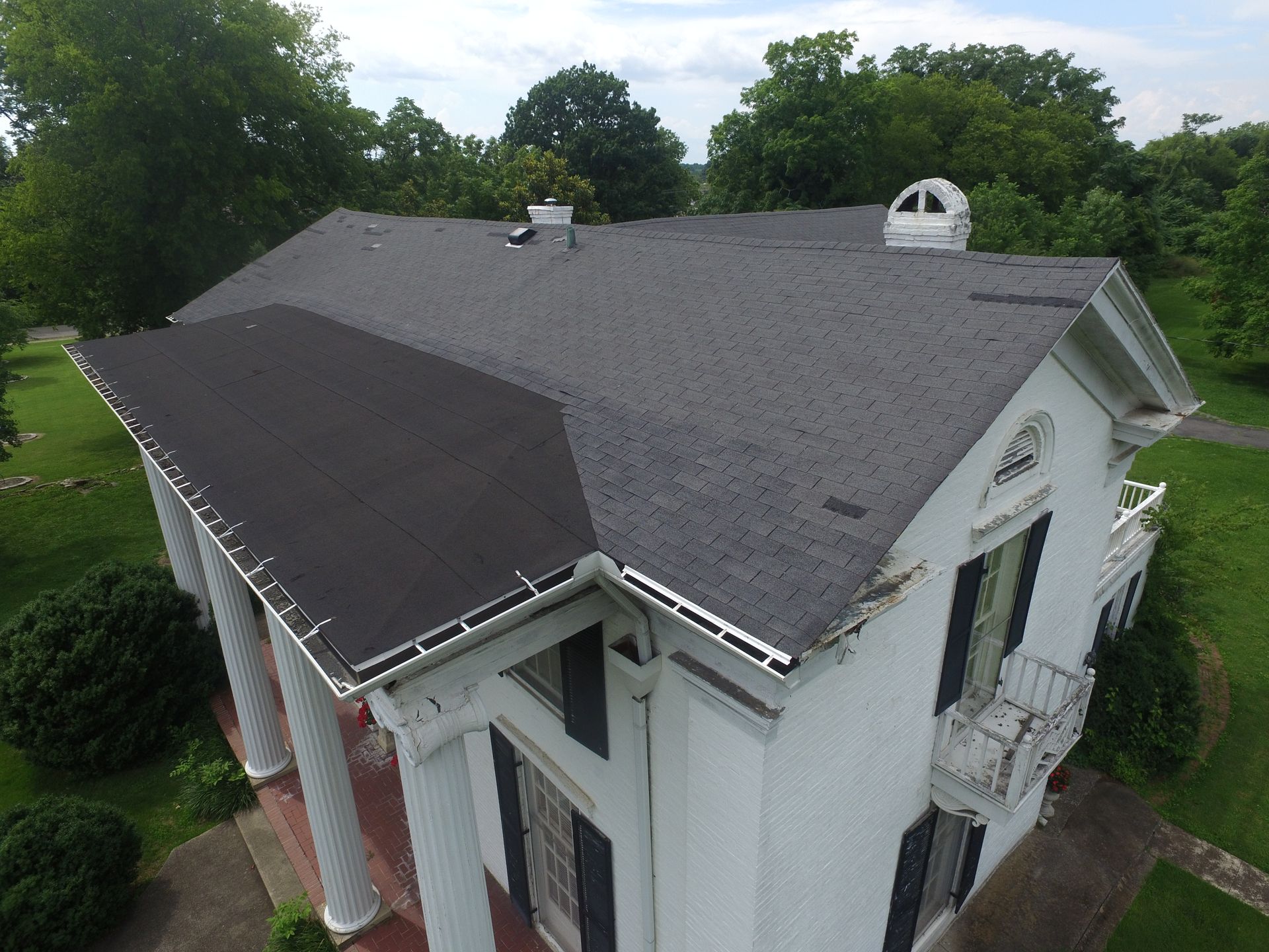 An elevated view of the shingled roof and white exterior of a classic, two-story house with columns and green trees.