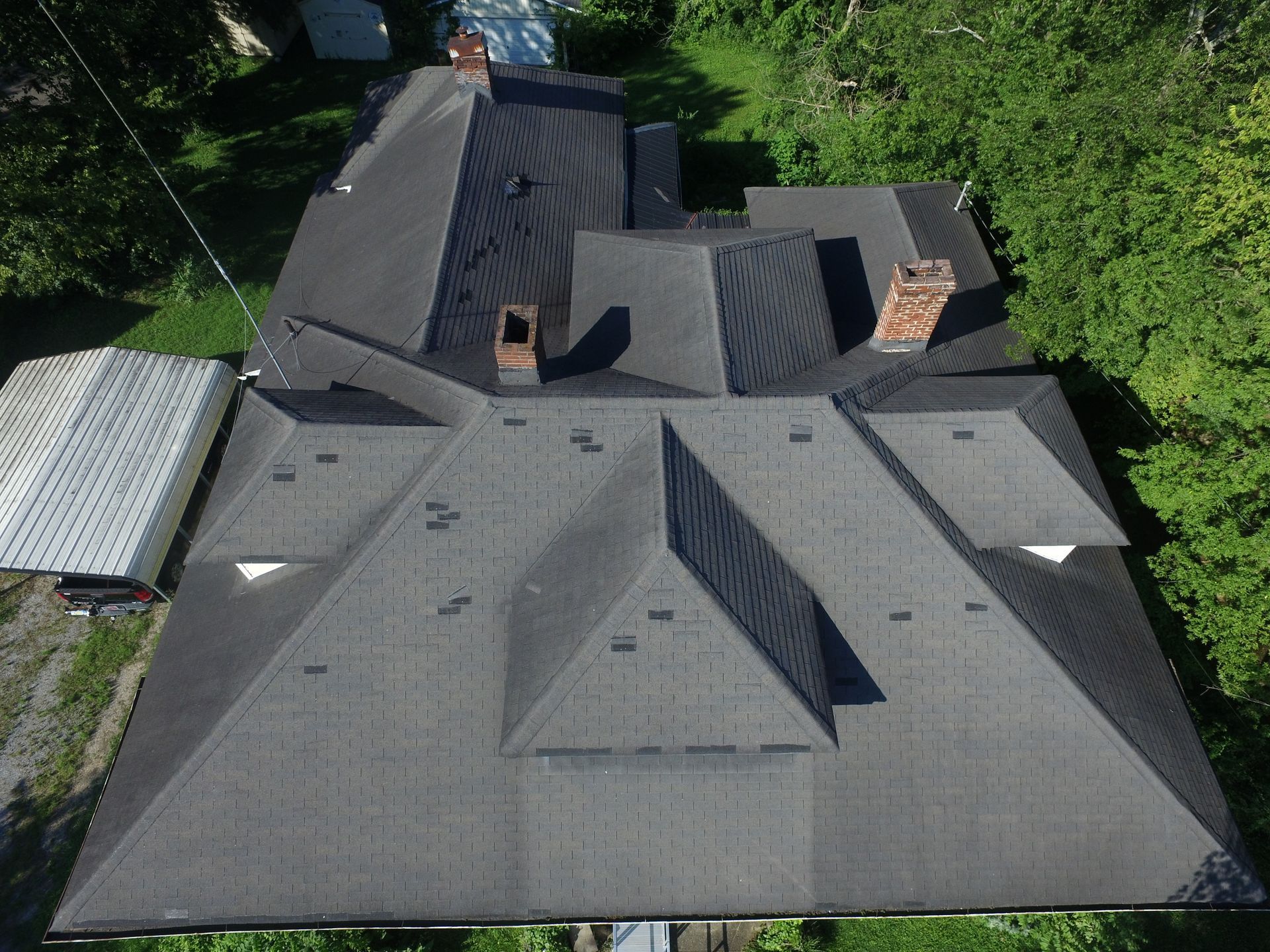 An aerial, top-down view of a complex, multi-faceted house roof with gray shingles, chimneys, and vents, surrounded by trees.