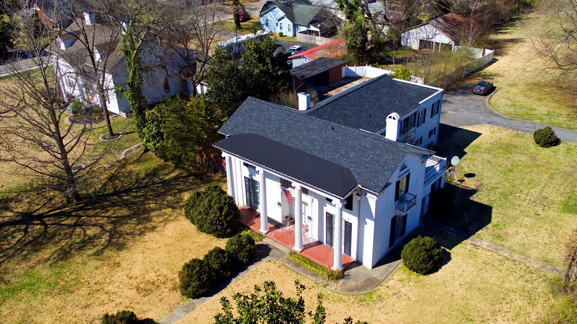 Aerial view of a white, two-story house with columns and a dark roof, surrounded by a lawn on a sunny day.