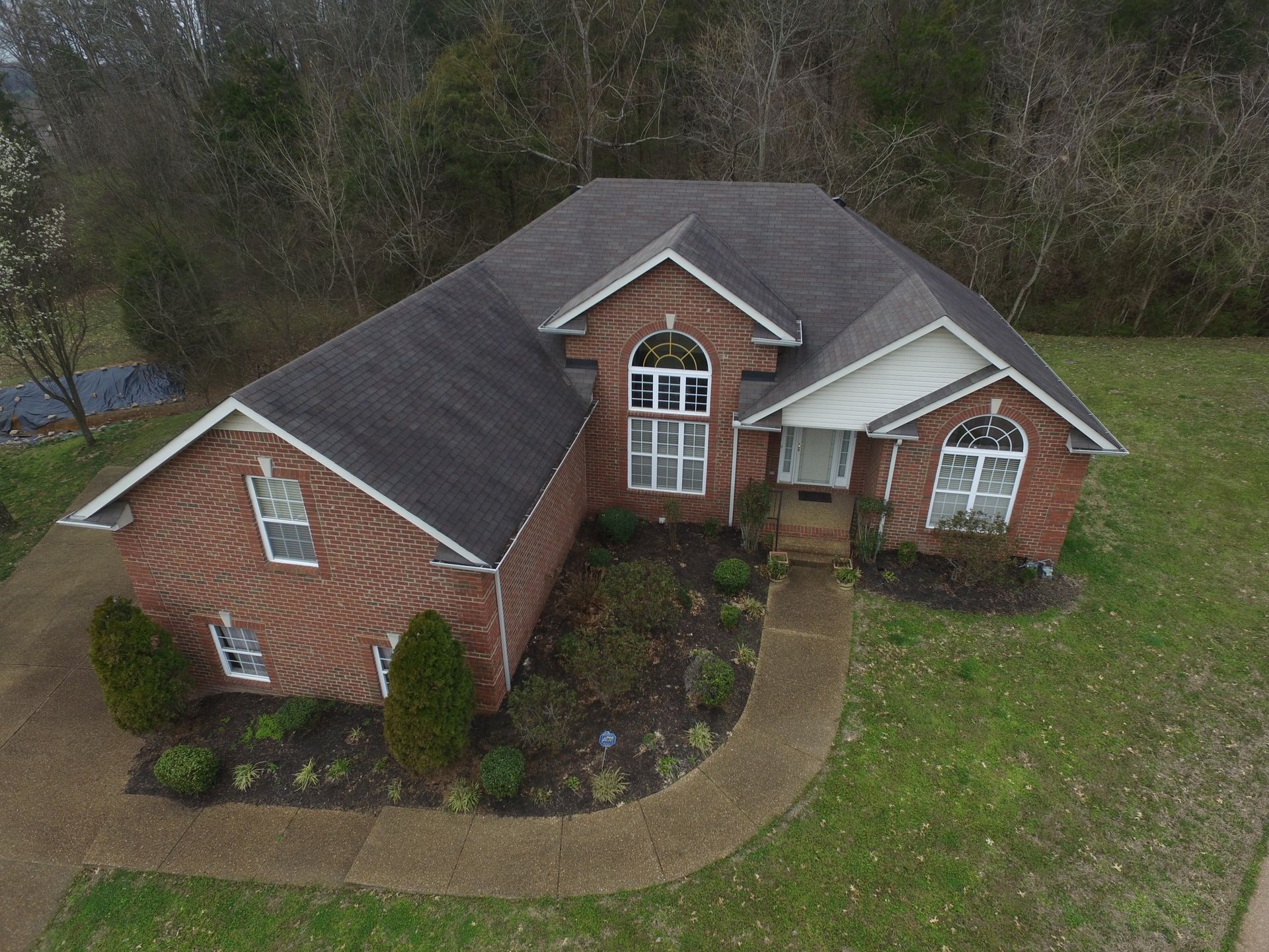 An aerial view of a single-story brick house with a dark shingled roof, a paved walkway, and a surrounding grass lawn.