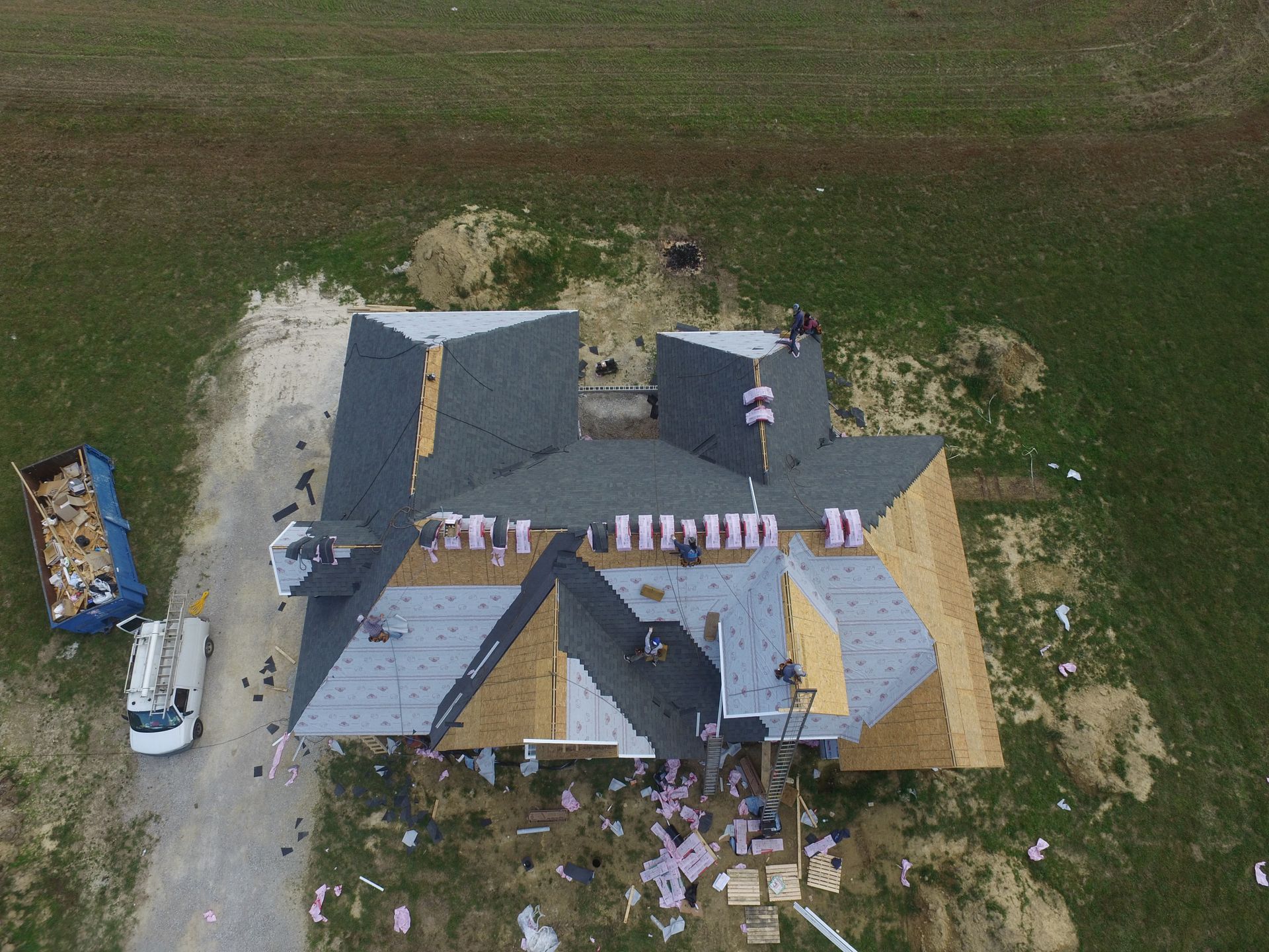 An aerial view shows workers on the roof of a house under construction, with materials, a dumpster, and a truck below.