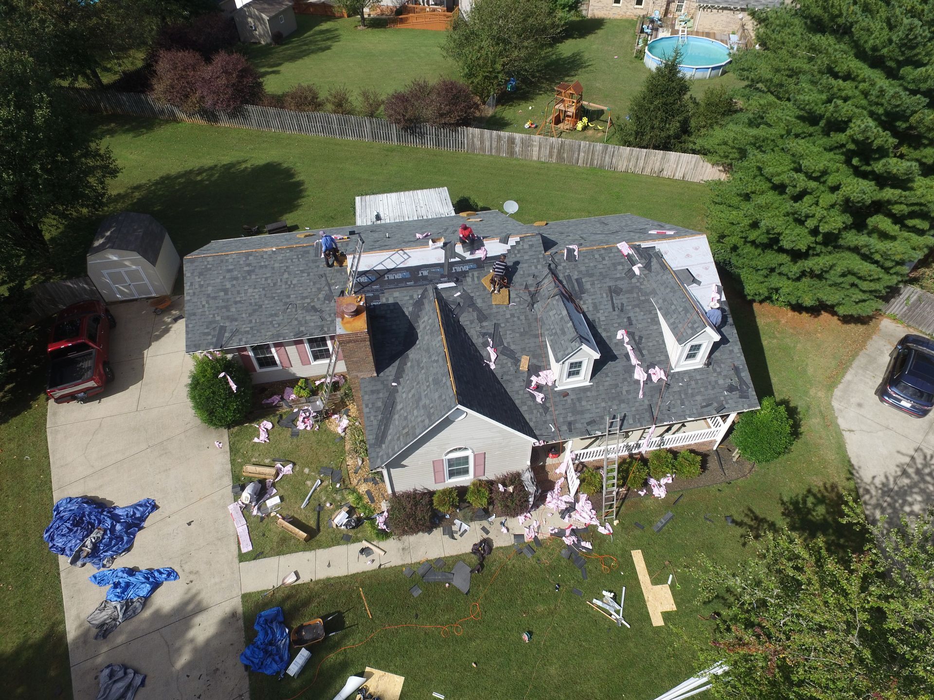 An aerial view of workers installing new grey shingles on a residential house roof with debris scattered on the lawn below.