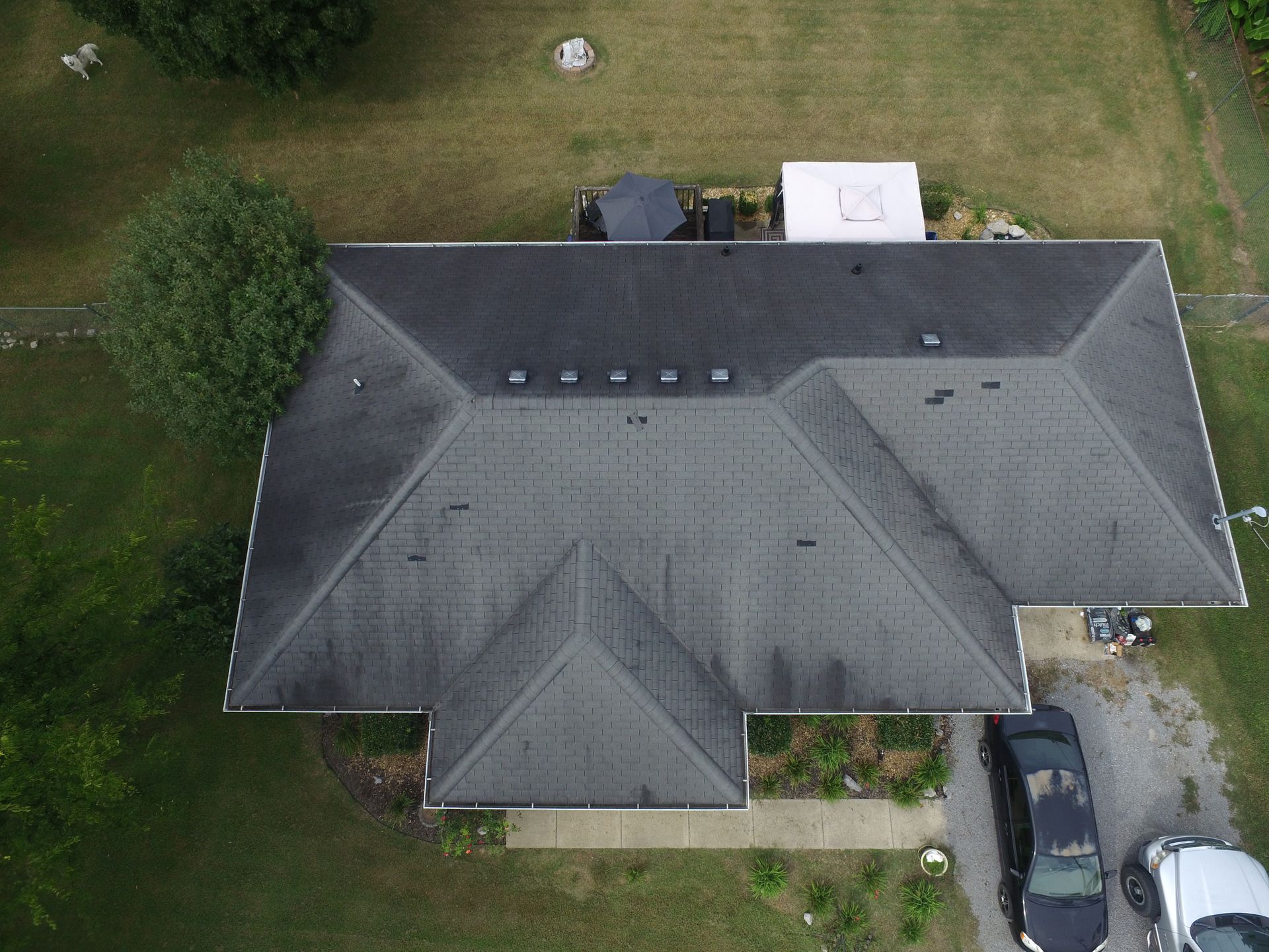 An aerial view of a rectangular house with a grey shingled roof, surrounded by green grass and two parked cars.
