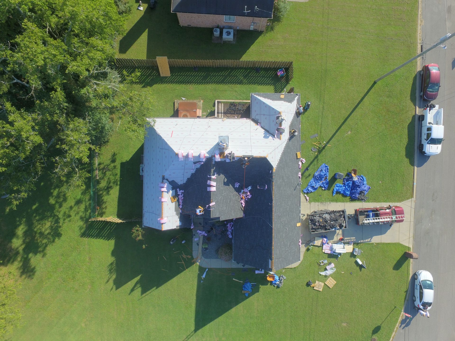 Aerial view of a residential roof replacement with workers, building materials, and tools on the roof and lawn.