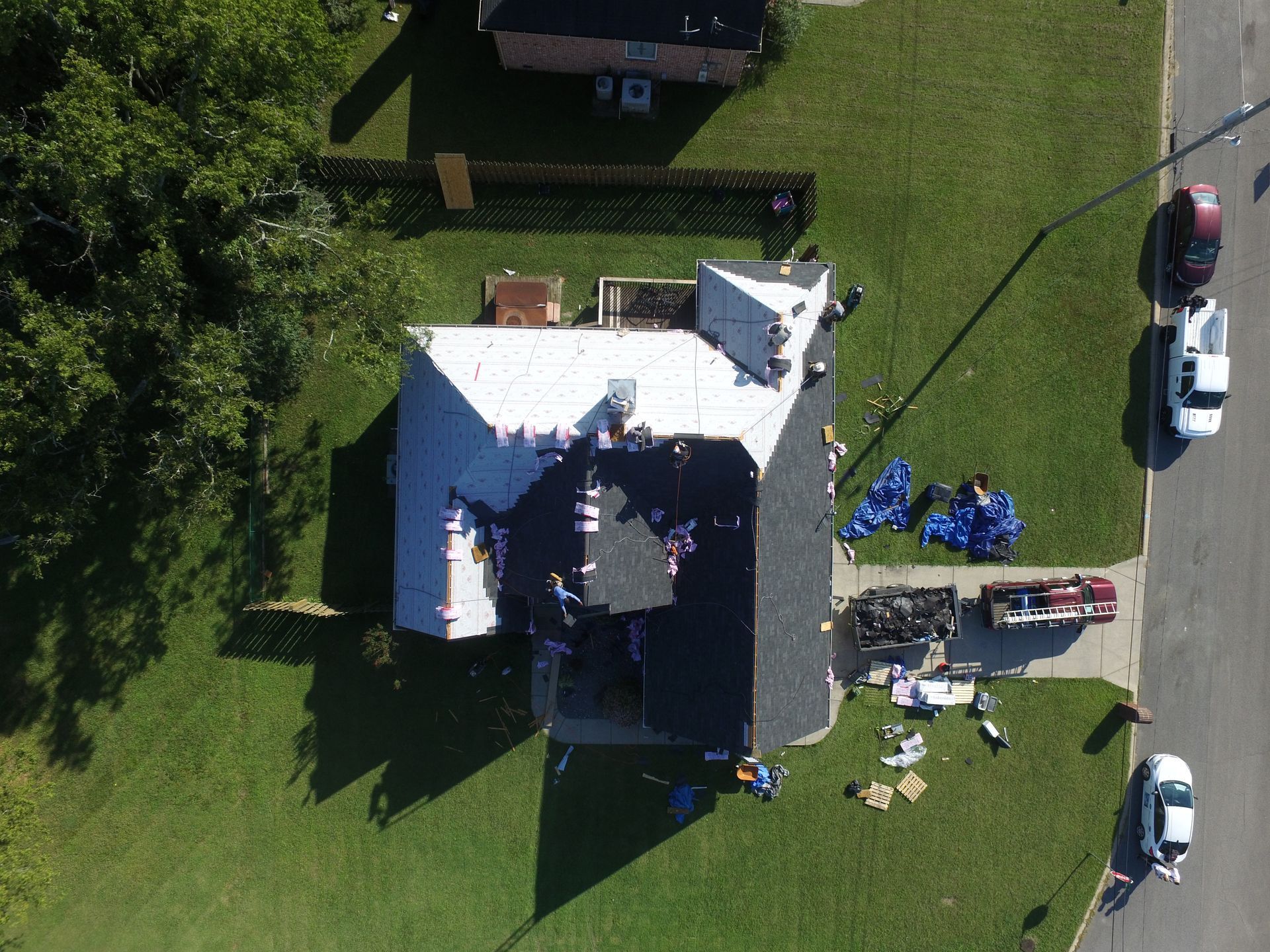 An aerial view of a house undergoing a roof replacement with workers, tools, and materials scattered on the roof and lawn.