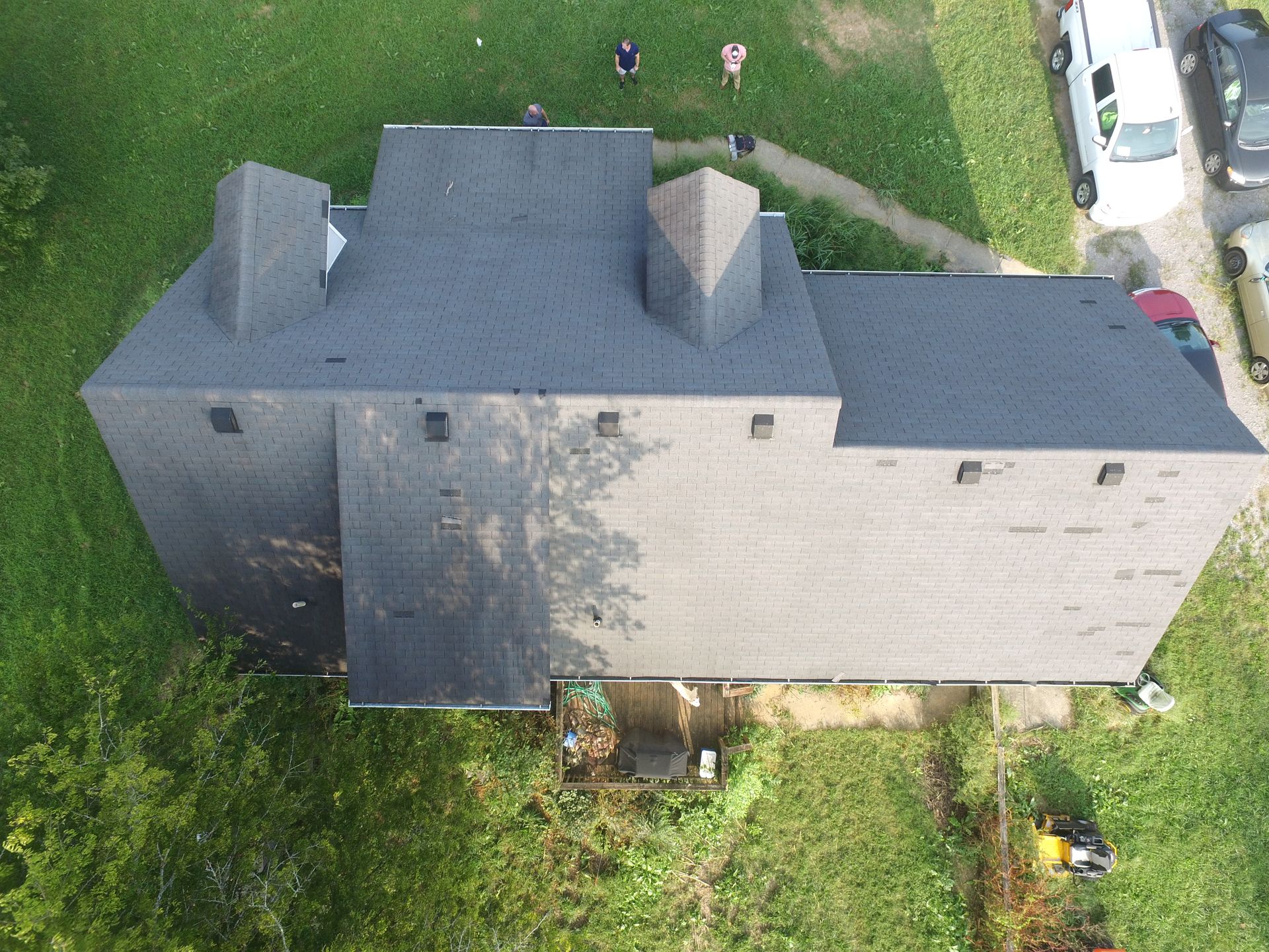 An overhead drone view of a shingled roof on a multi-story house, surrounded by green lawn, a driveway, and two people.