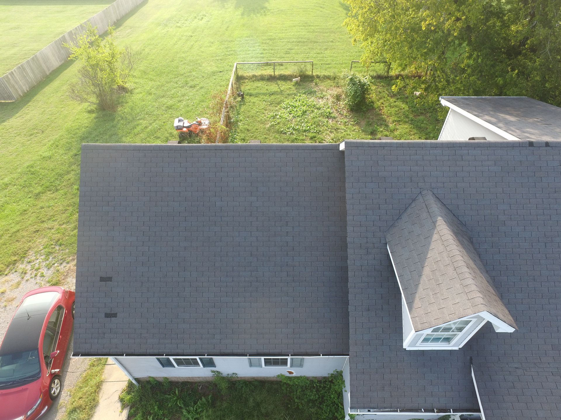 An aerial view of a gray shingled roof with a dormer, a red car parked nearby, and a grassy backyard.